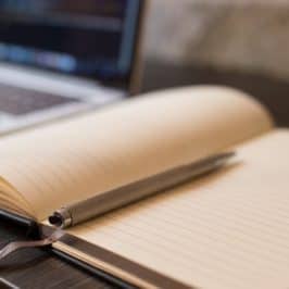 Open notebook with blank page, pen lying on top and laptop in background.