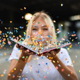 Woman blowing confetti off a book into the camera