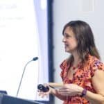 Woman with brown hair wearing orange blouse speaking at a podium.